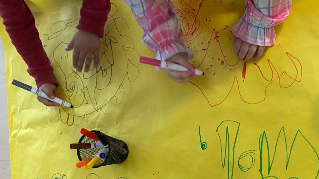Two young children draw colorful, abstract shapes and patterns on a large sheet of yellow paper using markers, with their hands and sleeves visible in the frame