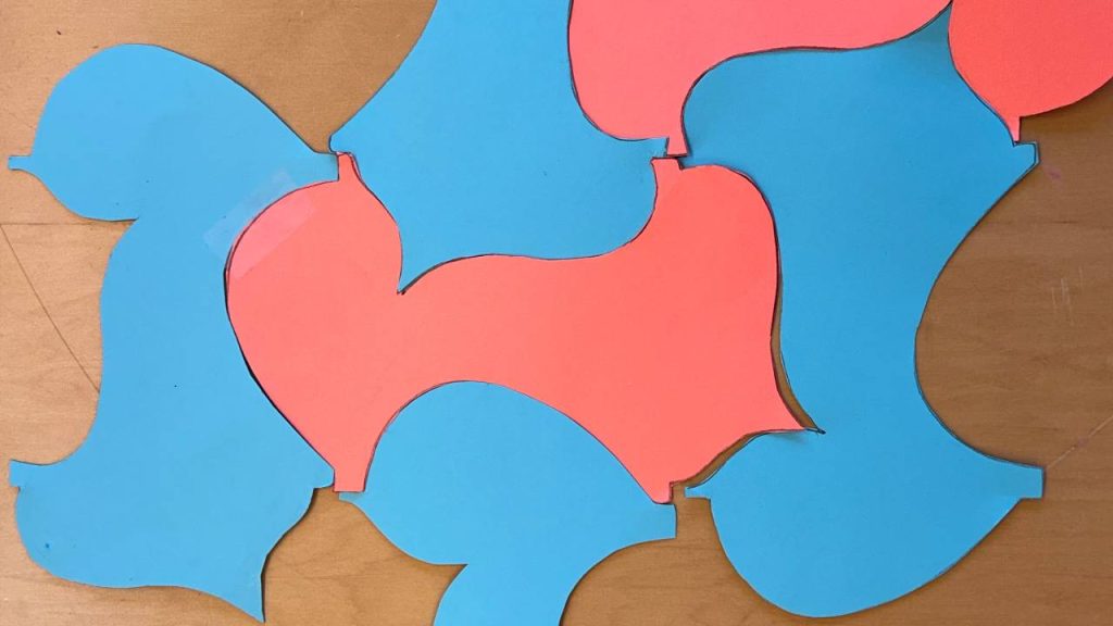 A student-created tessellation using interlocking pink and blue organic shapes cut from paper and arranged on a wooden surface.